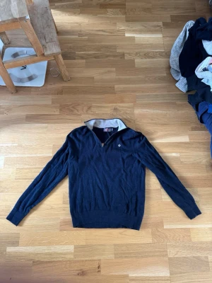 Marinblå quater ziptröja från Morris - Snygg marinblå ziptröja från Morris med halv dragkedja och kontrastfärgad krage i ljusgrått. Tröjan har en broderad logga på bröstet och ribbade muddar. Perfekt för en stilren och avslappnad look.