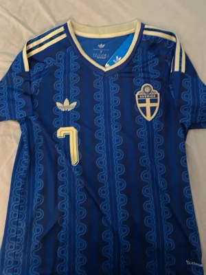 Sverige borta tröja fotboll bergvall - 📦FRAKTAR INOM 24H📦Säljer en blå fotbollströja från Adidas med Sveriges landslagsemblem och nummer 7 på bröstet. Tröjan har gulddetaljer, klassiska Adidas-ränder på axlarna och namnet 'Bergvall' på ryggen. Tillverkad i lätt och ventilerande material, perfekt för match eller träning.