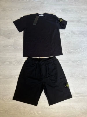 Svart sommar dress från Stone Island - Säljer ett par svarta shorts och T-shirt från Stone Island med klassiskt logopatch på benet och armen och snörning i midjan. Shortsen har en avslappnad passform och är perfekta för sommaren. Materialet känns mjukt och bekvämt, troligen bomull.