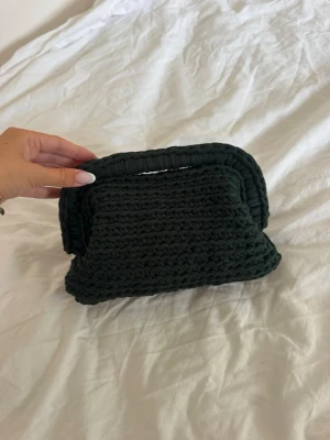 Mörkgrön virkad clutch - Unik handgjord clutch i mörkgrönt, virkat material. Väskan har en rektangulär form med integrerat handtag upptill, vilket ger en cool och trendig look. Perfekt accessoar för dig som vill sticka ut med något personligt och stilrent.