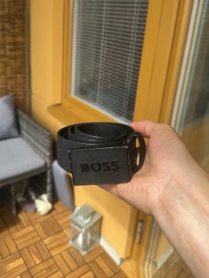 Svart BOSS bälte med metallspänne - Snyggt svart bälte från BOSS med stilren metallspänne där loggan är ingraverad. Bältet har en enkel och modern design som passar till de flesta outfits. Perfekt accessoar för dig som gillar klassiska detaljer.