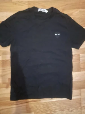 Svart t-shirt från Comme des Garçons Play - Svart t-shirt från Comme des Garçons Play med det ikoniska hjärta-loggan med ögon på bröstet. Klassisk passform och rund halsringning. Perfekt för en stilren och trendig look.