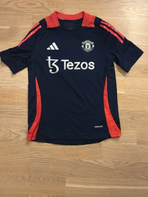 Manchester United tränings t-shirt Adidas - Snygg mörkblå Manchester United tränings t-shirt från Adidas med röda detaljer och logga på bröstet. Tillverkad i lätt och ventilerande material med Aeroready-teknologi. Perfekt för fotbollsträning eller sportiga dagar.