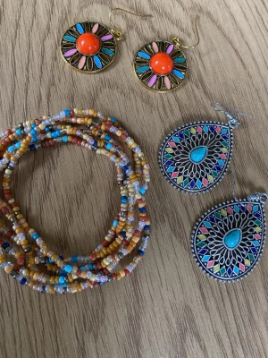Färgglada boho örhängen i metall - Säljer två par boho-inspirerade örhängen. Ett par är runda med guldfärgad metall, orange mitt och detaljer i turkos, rosa och beige. Det andra paret är droppformade i silverfärgad metall med turkos sten och färgglada mönster. Perfekta för dig som gillar statement-smycken.