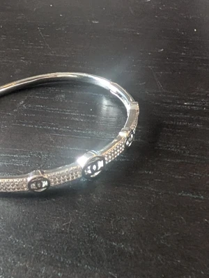 Silverfärgat CC armband med stenar - Snyggt silverfärgat CC armband med gnistrande små stenar längs hela bandet och flera ikoniska CC-loggor. Armbandet har en stilren och elegant design som verkligen sticker ut på handleden. Perfekt accessoar för dig som gillar exklusiva detaljer.