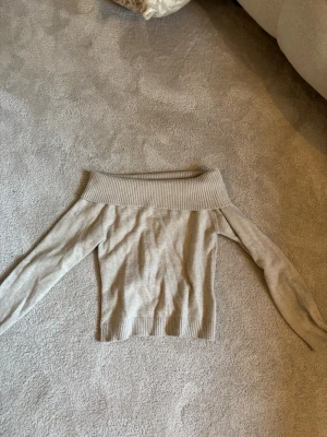 Beige off shoulder stickad tröja - Säljer en beige stickad off shoulder-tröja med bred ribbad kant upptill. Tröjan har långa ärmar och är gjord i mjukt material, perfekt för en trendig och avslappnad look. Passar till både jeans och kjol.