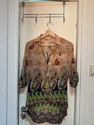 Mönstrad skjorta i tunnt material - Säljer en färgglad skjorta med paisley- och blommönster i beige, grön, svart och orange. Skjortan är tillverkad i ett tunt, luftigt material och har knappar framtill samt uppvikbara ärmar med knapp. Perfekt för dig som gillar unika plagg!