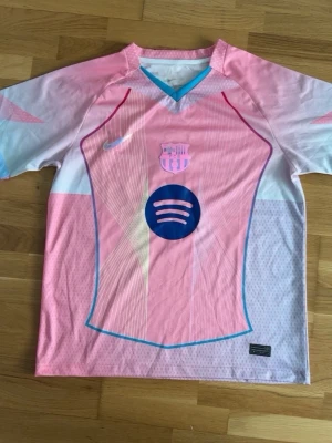 Rosa FC Barcelona matchtröja Nike - Snygg rosa FC Barcelona matchtröja från Nike med klubbmärke och Spotify-logga på bröstet. Tröjan har vita och ljusblå detaljer, V-ringning med blå kant och är tillverkad i ett lätt, ventilerande funktionsmaterial. Perfekt för fotboll eller träning.