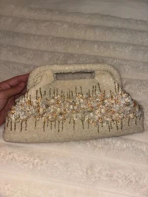 Zara Pärlbroderad vit kuvertväska - Säljer en unik vit kuvertväska med inbyggt handtag. Väskan är täckt av små pärlor och har ett detaljerat broderi med vita och guldiga pärlor samt paljetter i mitten. Perfekt accessoar för att lyfta din outfit med en elegant touch.