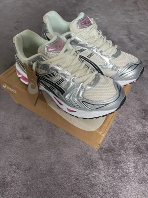Asics gel kayano 14  - Snygga sneakers från Asics i silver och beige mesh med svarta och rosa detaljer. Skorna har en chunky sula med rosa inslag och klassisk Asics-logga på sidan. Ovandelen är i mesh och syntet för en sportig vibe. Perfekta för dig som gillar retrostil och streetwear.