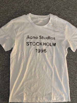 Vit Acne Studios t-shirt Stockholm - Säljer en vit t-shirt från Acne Studios med trycket 'STOCKHOLM 1996' på bröstet. Klassisk rund hals och korta ärmar. Tillverkad i mjuk bomull för en skön och avslappnad look.