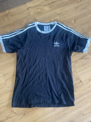 Svart Adidas t-shirt med vita detaljer - Klassisk svart t-shirt från Adidas med vita ränder. Använd ett fåtal gånger och bara legat