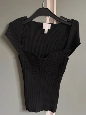 Svart ribbad topp från H&M - Säljer en svart ribbad topp från H&M med markerad byst och breda axelband. Toppen har en tight passform och är tillverkad i stretchigt material som sitter snyggt på kroppen. Perfekt att styla med jeans eller kjol.