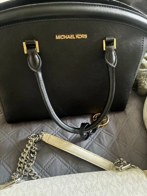 Svart handväska från Michael Kors - Stilren svart handväska från Michael Kors i strukturerat läder med guldfärgade detaljer och dubbla handtag. Väskan har en klassisk bågformad design och logotypen i guld framtill. Perfekt för dig som gillar en elegant och tidlös look.