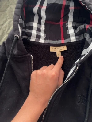 Svart hoodie från Burberry med dragkedja - Svart hoodie från Burberry med klassiskt rutigt foder i huvan och dragkedja framtill. Broderad logga på bröstet och justerbar dragsko i huvan. Mjuk insida och snygga metalländar på snörena. Perfekt för en stilren och bekväm look.