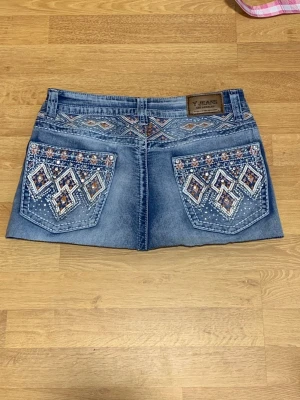 Broderad jeanskjol med mönster - Kort jeanskjol från Y Jeans i ljusblå denim med stora broderade bakfickor i vitt och brunt. Kjolen har klassisk femficksdesign, knapp och dragkedja framtill samt dekorativa detaljer på fickorna. Perfekt för dig som gillar unika detaljer och denim.