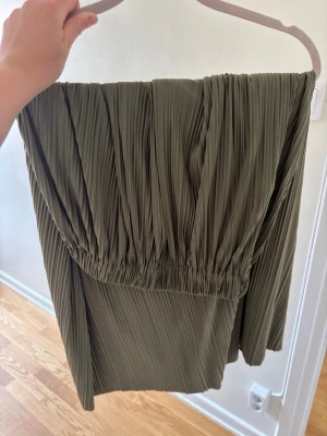 Plisserad grön offshoulder klänning - Trendig kortklänning i plisserat tyg med offshoulder-design och enarmad ärm. Klänningen är olivgrön och har en loose passform som ger en cool och avslappnad vibe. Perfekt för dig som gillar unika detaljer och stilren look.
