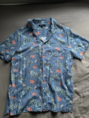 Skjorta - Säljer en kortärmad blå skjorta från Mywearman med blommigt mönster i vitt, grönt och orange. Skjortan har klassisk krage och knappar framtill. Perfekt för sommarens alla tillfällen!