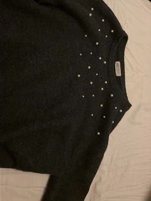 Mörkgrå stickad tröja med pärlor H&M - Mysig mörkgrå stickad tröja från H&M med dekorativa pärlor över axlar och bröst. Tröjan har rund halsringning och långa ärmar. Perfekt för dig som vill ha en enkel men ändå trendig look.