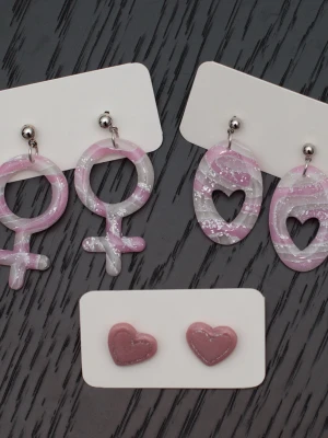 Handgjordas örhängen i polymerlera och stift i rostfritt stål feminist kvinnosymbol rosa - Handgjorda örhängen i polymerlera och stift i rostfritt stål.  Självklart nya och oanvända. Storlek: Ovala med hjärtan 2x4cm. Kvinnosymbol 2,5x5cm. Stiftörhängen 1,5x1,5cm.  Färgerna kan variera beroende på skärminställningar.