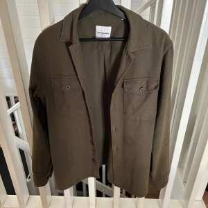 Grön overshirt från Jack & Jones - Stilren grön overshirt från Jack & Jones med klassisk krage och två bröstfickor med knappar. Skjortan har långa ärmar med dubbla manschettknappar och är tillverkad i mjukt bomullsmaterial. Perfekt att bära som lager över en t-shirt för en chill look. En manschettknapp är sönder på ena ärmen annars inga skador.
