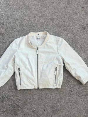 Vit bomberjacka från Iina med Trekvarts ärm - Snygg vit bomberjacka från Iina med klassisk rund krage och dragkedja framtill. Jackan har två framfickor med dragkedjor och detaljer i samma färg. Materialet ser ut att vara mjukt och lätt, perfekt för en trendig look.