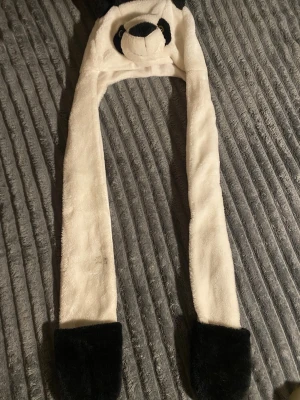 Panda mössa med långa öronlappar - Söt och mjuk panda mössa i vitt och svart. Mössan har långa öronlappar som man kan ha händerna i och ett broderat pandatryne med svarta öron och ögon. Perfekt för dig som vill sticka ut med en rolig accessoar.
