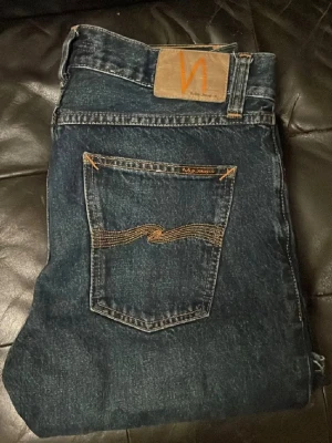 Mörkblå jeans från Nudie Jeans - Säljer ett par klassiska mörkblå jeans från Nudie Jeans med raka ben och snygga kontrastsömmar. Jeansen har en ikonisk bakficka med vågig söm och patch i läder baktill i midjan. Tillverkade i slitstark denim, perfekta för en avslappnad och stilren look. Storleken är 33/34