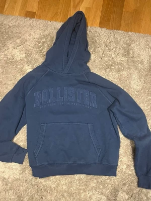 Blå hoodie från Hollister - Mörkblå hoodie från Hollister med stor huva och magficka. Broderad logga på bröstet i samma ton som tyget. Klassisk passform med ribbade muddar vid ärmar och nederkant. Tillverkad i mjuk bomullsblandning för extra komfort.
