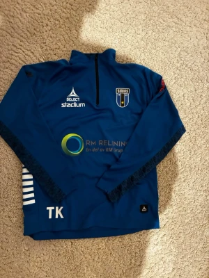 Blå Sirius träningströja Select - Säljer en blå långärmad träningströja från Sirius med halv dragkedja framtill. Tröjan har tryckta loggor från Select, Stadium och flera sponsorer samt klubbmärke på bröstet. Snygga vita detaljer på sidorna och initialerna TK nedtill.