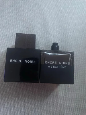 Encre Noire parfym duo - Två flaskor Encre Noire parfym från Lalique. Båda flaskorna är svarta med minimalistisk design och vit text. En har träliknande lock och den andra är Encre Noire À L’Extrême med spraymunstycke. Flaskorna är fyrkantiga och stilrena.