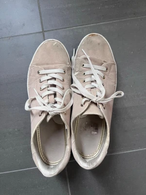 Beige sneakers med vita snören - Säljer ett par beige sneakers från XTI med klassisk design och vita snören. Skorna har en låg profil och vit sula med mönster för extra grepp. Materialet är textil och syntet, perfekt för en avslappnad och stilren look.