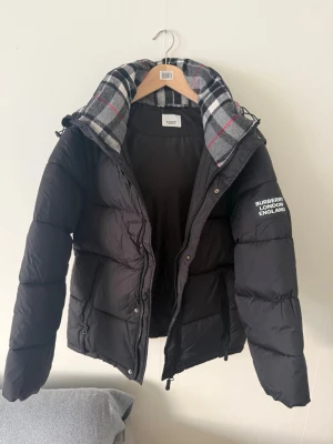 Svart pufferjacka från Burberry - Stilren svart pufferjacka från Burberry med klassiskt rutigt foder i huvan och tydlig logga på ärmen. Jackan har dragkedja, hög krage och en oversized look som ger en cool vibe. Perfekt för kalla dagar när du vill hålla stilen. Du kan även ha den som en väst perfekt för sommaren eller våren!  PRIS GÅR ATT DISKUTERAS