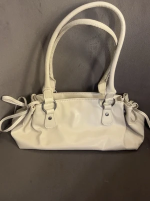 Vit handväska med knytdetaljer - Stilren vit handväska i slätt konstläder med dubbla handtag och snygga knytdetaljer på sidorna. Väskan har en rektangulär form och svart innerfoder