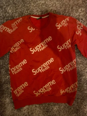 Röd Supreme Paris sweatshirt - Röd sweatshirt från Supreme Paris med vitt logotryck över hela tröjan. Rund halsringning och ribbade muddar vid ärmslut och nederkant. Perfekt för dig som gillar streetwear och vill sticka ut med en ikonisk design.