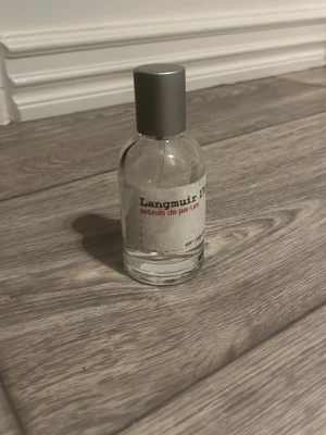 Langmuir Extrait de Parfum - En stilren parfymflaska från Langmuir med transparent glas och en enkel, rundad form. Flaskan har en grå metallfärgad kork och en vit etikett med svart och röd text. Perfekt för dig som gillar minimalistisk design.
