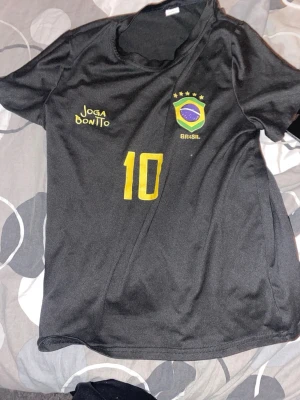 Svart Brasilien fotbollströja #10 - Snygg svart fotbollströja med Brasilien-emblem och nummer 10 i gult på framsidan. Trycket 'Joga Bonito' på bröstet och klassisk rund hals. Tillverkad i lätt och ventilerande material, perfekt för träning eller match.