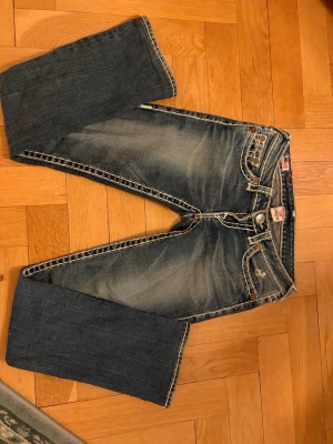 True Religion bootcut jeans blå - Säljer ett par blå bootcut jeans från True Religion med tydliga kontrastsömmar och ikoniska broderade fickor bak. Jeansen har låg midja och slitningar framtill för en cool, avslappnad look. Klassisk femficksmodell i bomull. Jeansen är lite slimmade vid vaderna, så inte riktigt bootcut.