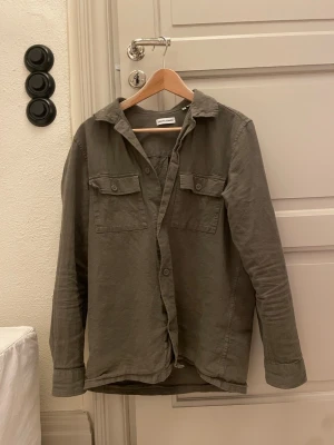Olivgrön overshirt  - Snygg olivgrön skjorta/overshirt från Jack & Jones med två bröstfickor och knappar framtill. Skjortan har klassisk krage och lång ärm, tillverkad i mjuk bomull för en avslappnad look.