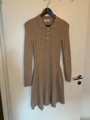 Beige ribbad klänning Holly & Whyte - Snygg beige ribbad stretchig klänning från Holly & Whyte med klassisk krage och fyra guldfärgade knappar framtill. Klänningen har lång ärm och är figurnära upptill med en lätt utsvängd kjol. Perfekt för dig som gillar stilrena och bekväma plagg.