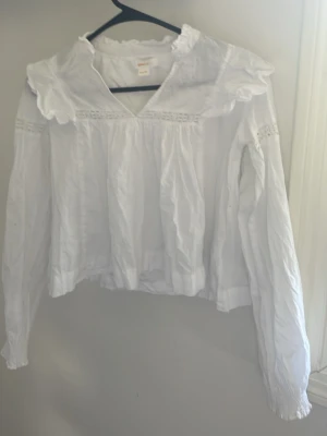 Vit blus med volangkrage - Söt vit blus med lång ärm och volangkrage. Blusen har v-ringning och dekorativa spetsdetaljer vid axlarna. Tillverkad i ett lätt tyg som ger en romantisk och luftig känsla. Perfekt för dig som gillar en feminin och trendig stil.