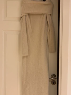 Beige offshoulder stickad klänning - Mysig beige stickad klänning med offshoulder-design och långa ärmar. Klänningen har en rak passform och är perfekt för dig som gillar en enkel men ändå trendig look. Materialet är mjukt och värmande, vilket gör den skön att bära under kyligare dagar.