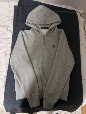 Polo Ralph Lauren hoodie - Snygg grå hoodie med dragkedja från Polo Ralph Lauren. Klassisk design med huva, två fickor fram och den ikoniska logon broderad på bröstet. Perfekt för chill dagar och enkel att matcha med jeans eller joggers. Materialet är mjukt och bekvämt.