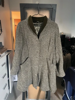 Gråemerald rock från VIC Ulla Hannerz - Stilren gråemerald rock från VIC Ulla Hannerz med bred krage i mörkare tyg och stora fickor framtill. Rocken har en avslappnad passform och knäppning framtill. Materialet är grovt vävt och fodret är slätt och grått.