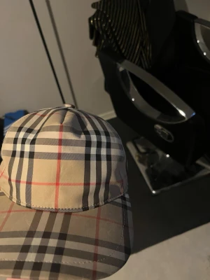 Beige rutig keps från Burberry - Säljer en klassisk beige keps från Burberry med det ikoniska rutiga mönstret i svart, vitt och rött. Kepsen har böjd skärm och justerbar rem bak. Perfekt accessoar för dig som vill ha en tidlös och trendig look.