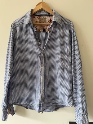 Blå rutig skjorta   - Snygg blårandig skjorta från Scotch & Soda med klassisk krage och knäppning framtill. Skjortan har en cool detalj på högersidan och blommigt innerfoder i kragen. Har en liknande i rött också, styck 200 men tillsammans 250 kr.