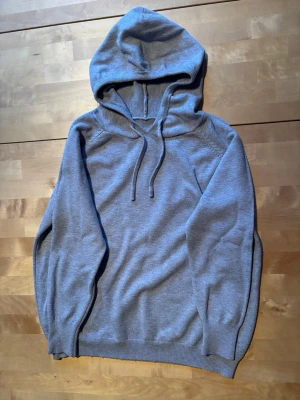 Grå basic hoodie med dragsko - En stilren grå hoodie med luva och dragsko. Tillverkad i mjukt, tunt stickat material som känns skönt mot huden. Klassisk passform med ribbade muddar vid ärmslut och nederkant. Perfekt för en avslappnad look.