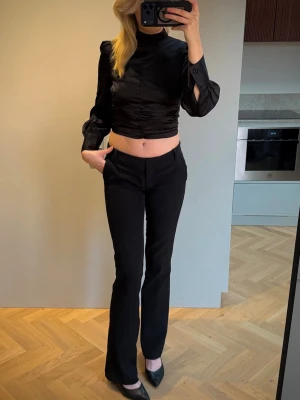 Svart blus med knytning - Svart croppad blus med långa ärmar och hög krage. Blusen har ett knytband i ryggen och breda manschetter vid ärmsluten.