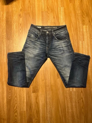 Blå jeans från Jack & Jones - Säljer ett par snygga blå jeans från Jack & Jones med slitningar och tvättade detaljer. Klassisk femficksmodell med normal midja och raka ben. Jeansen har en cool faded effekt och är tillverkade i denim med stretch för extra komfort.