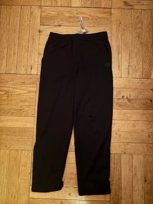 Svarta mjukisbyxor från Acne Studios - S/M Svarta mjukisbyxor från Acne Studios med klassisk rak passform och elastisk midja. Byxorna har den ikoniska Acne Studios-loggan med ansikte på vänster ben. Tillverkade i mjukt bomullsmaterial för maximal komfort.